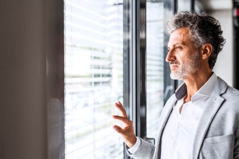 A man with gray hair and a beard looks out of a window with blinds, deep in thought as he contemplates finalizing his divorce. Wearing a light gray suit and white shirt, sunlight filters through the blinds, casting stripes of light and shadow on his face.