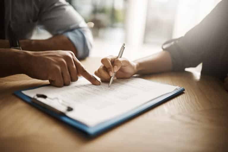 Two people reviewing a document on a clipboard at a table. One points to the paper while the other, holding a pen, seems ready to respond to divorce papers. The setting is well-lit, with a focus on the hands and document, suggesting an important discussion or signing process.