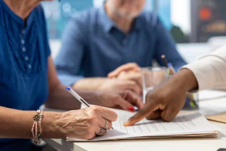 Two people are sitting at a table reviewing documents, focusing on important clauses. One person holds a pen, signing a paper, while the other points out specific sections of the postnuptial agreement. Both are in casual clothing against a blurred background, where another person is slightly visible.