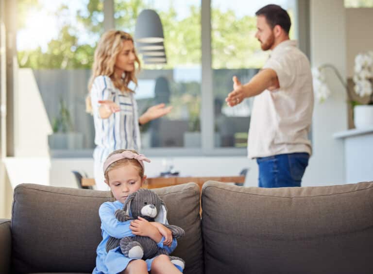 A young girl clutches a stuffed toy on the couch with a concerned expression. In the background, a couple argues about child support, their hands raised in frustration. The room is bright with large windows and modern decor, typical of an Oregon home.