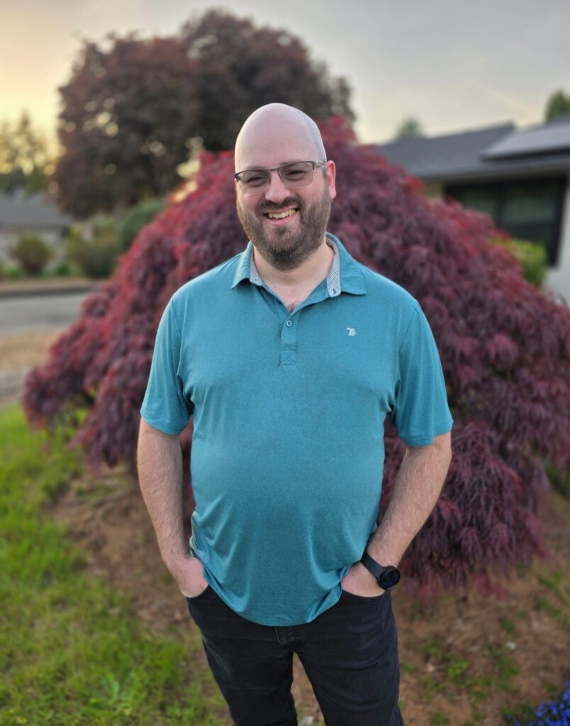 Joseph Crouch, a man with glasses and a beard, smiles at the camera while standing outdoors in front of a burgundy-leaved shrub. He wears a teal polo shirt, black pants, and a watch, with his hands in his pockets.
