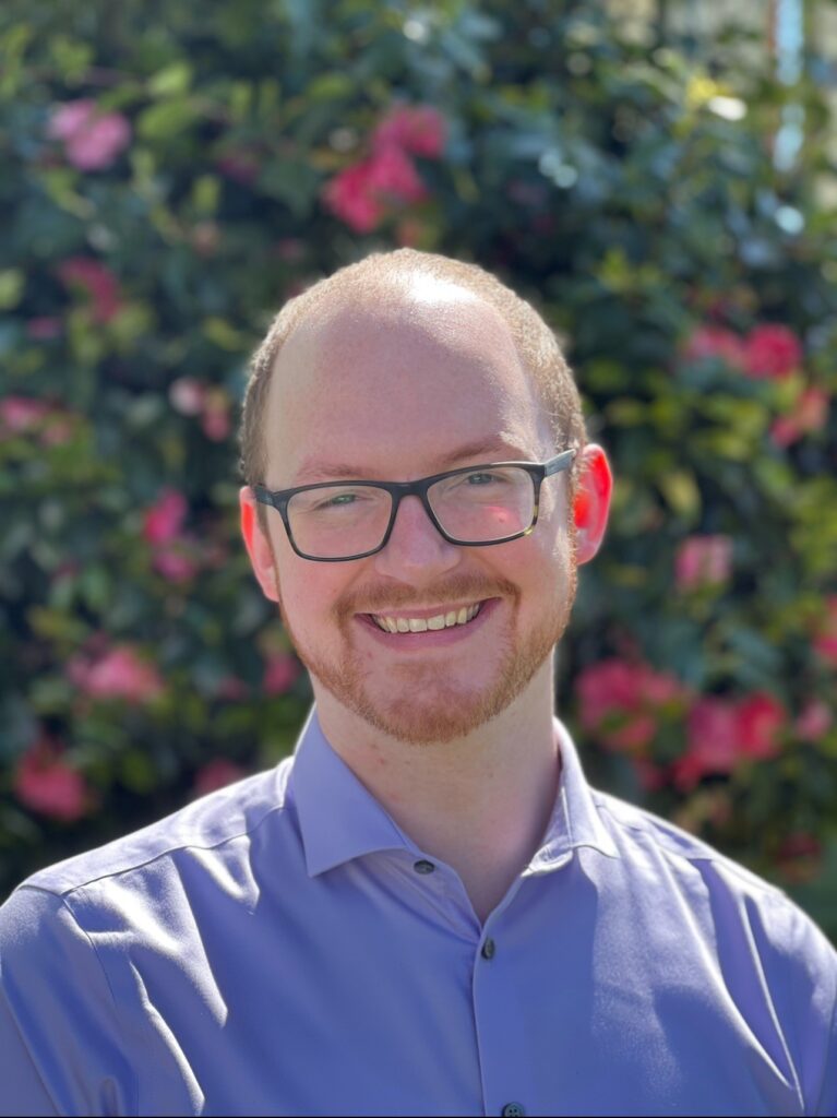 Joseph Crouch, a smiling person with glasses, a beard, and short hair wearing a light purple shirt, stands outdoors in front of green foliage with pink flowers, bathed in sunlight.